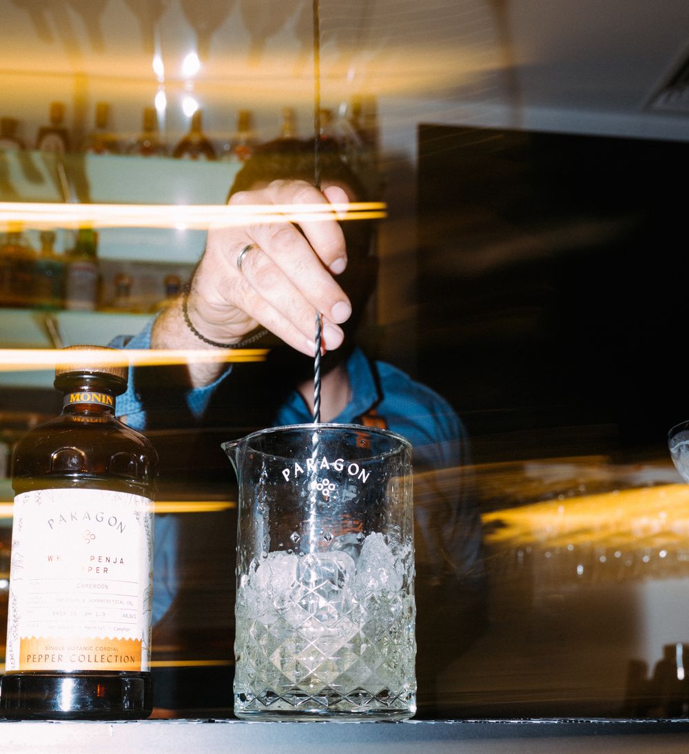 A bartender pours a cocktail into a cut crystal mixing glass with ice at a dimly lit bar.