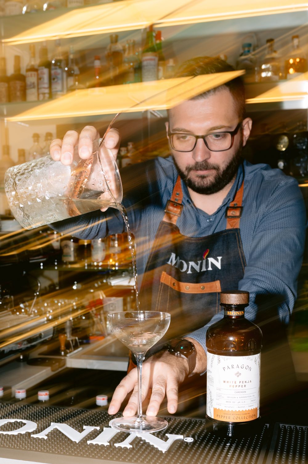 A bartender in an apron pours a cocktail at a bar with bottles displayed on shelves in the background.