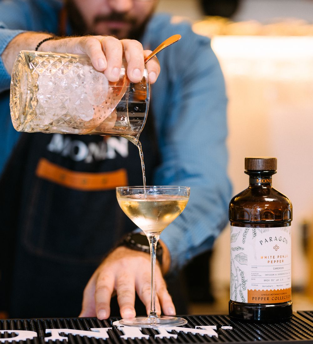 A bartender pours a cocktail from a crystal mixing glass into a coupe glass next to a dark bottle of spirits.