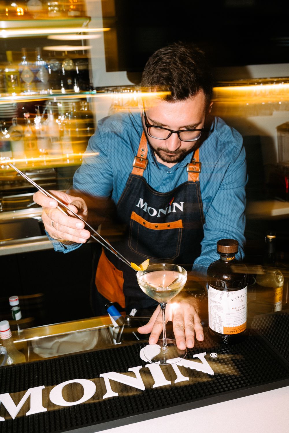 A bartender in a brown apron carefully garnishes a cocktail at a Monin-branded bar counter with bottles in the background.