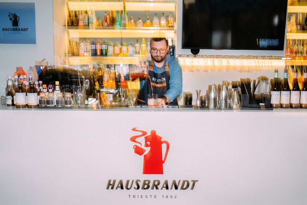 Bartender standing behind a modern white bar counter with a red Hausbrandt logo and various bottles displayed.