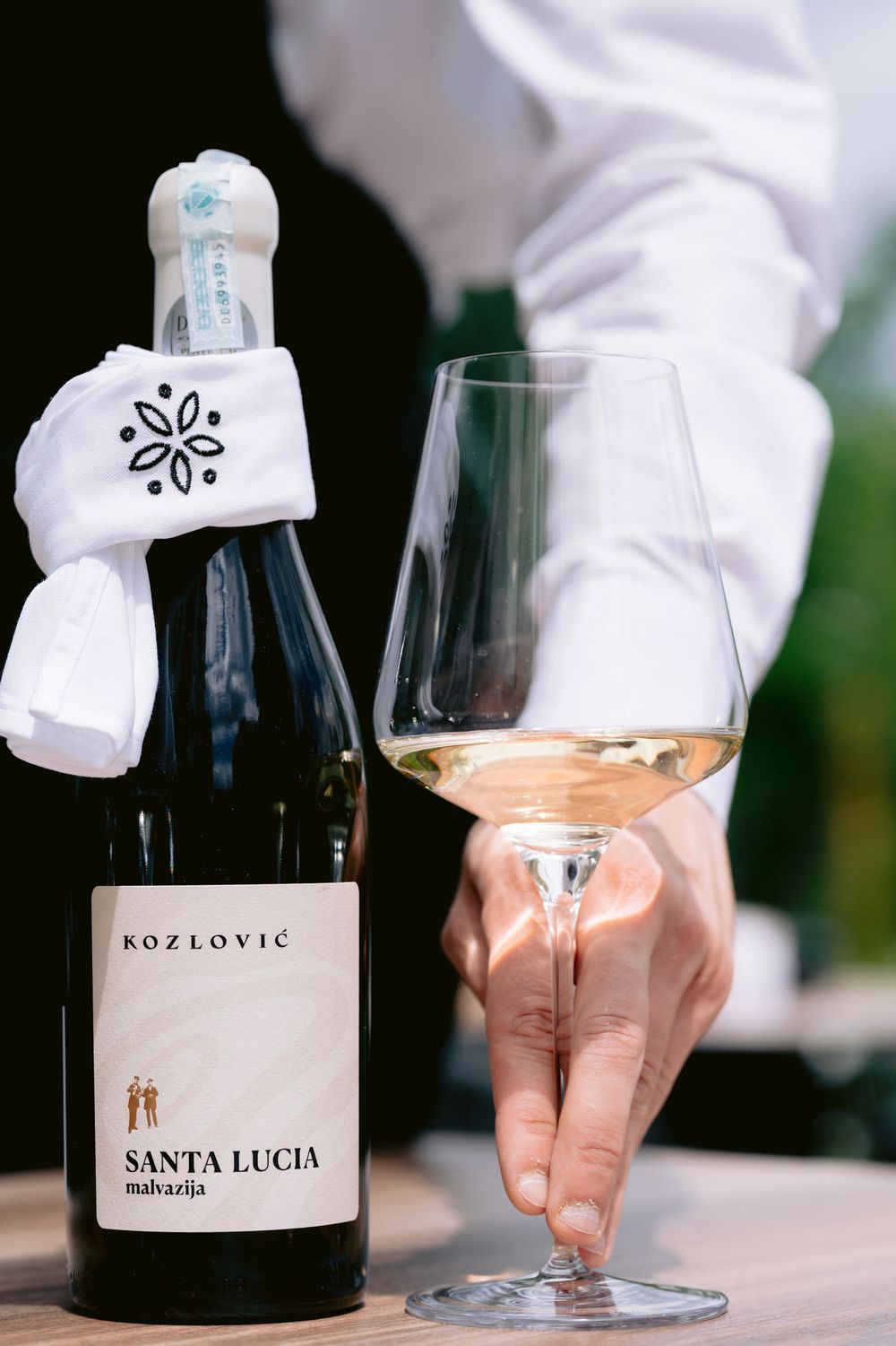 A bottle of Kozlovic Santa Lucia wine sits next to a glass of rose with a decorative white bottle wrap on a wooden table.