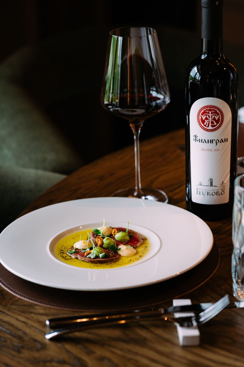 A series of wine bottles and elegant plated dishes with yellow soup is displayed on a dark wooden table in moody lighting.