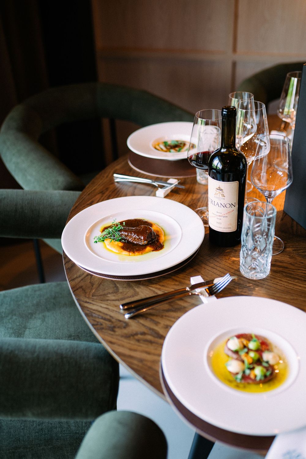 Elegant restaurant table setting with three white plates of gourmet dishes and a bottle of red wine beside green velvet chairs.