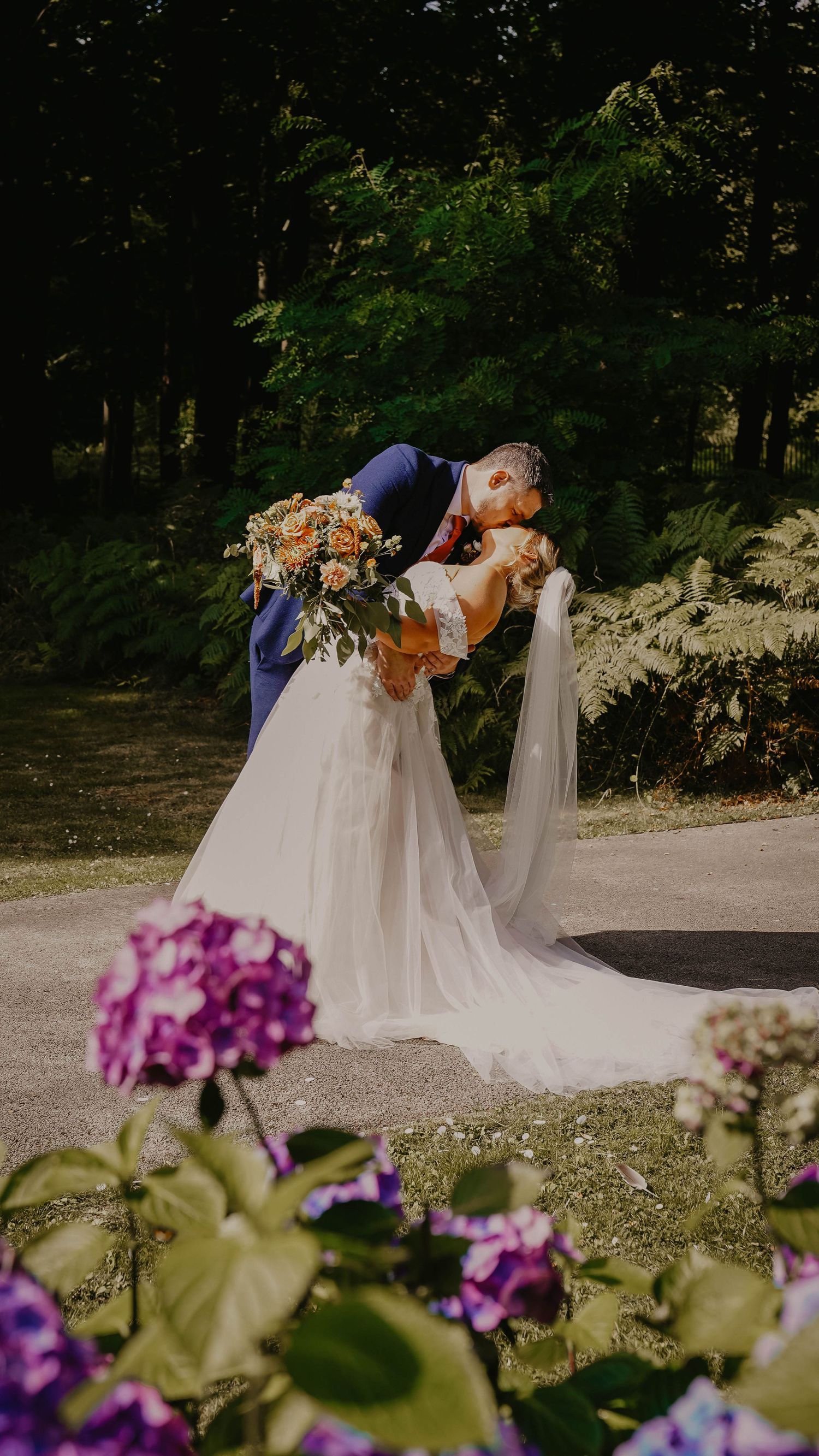 Romantic dip kiss moment in garden setting with purple flowers in foreground.
