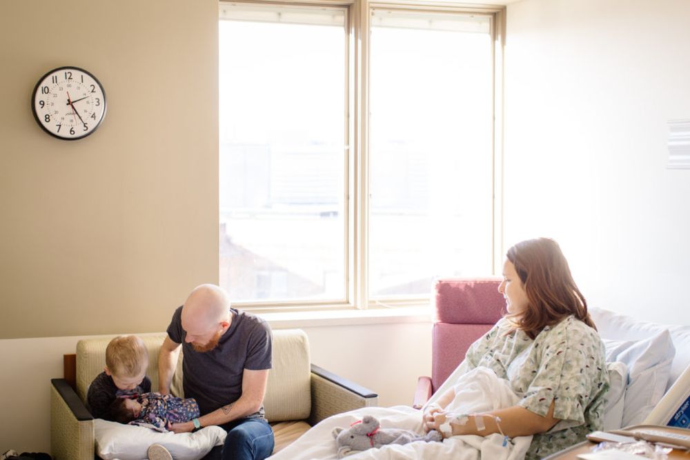 Family spending time together in hospital room with newborn and medical equipment visible.
