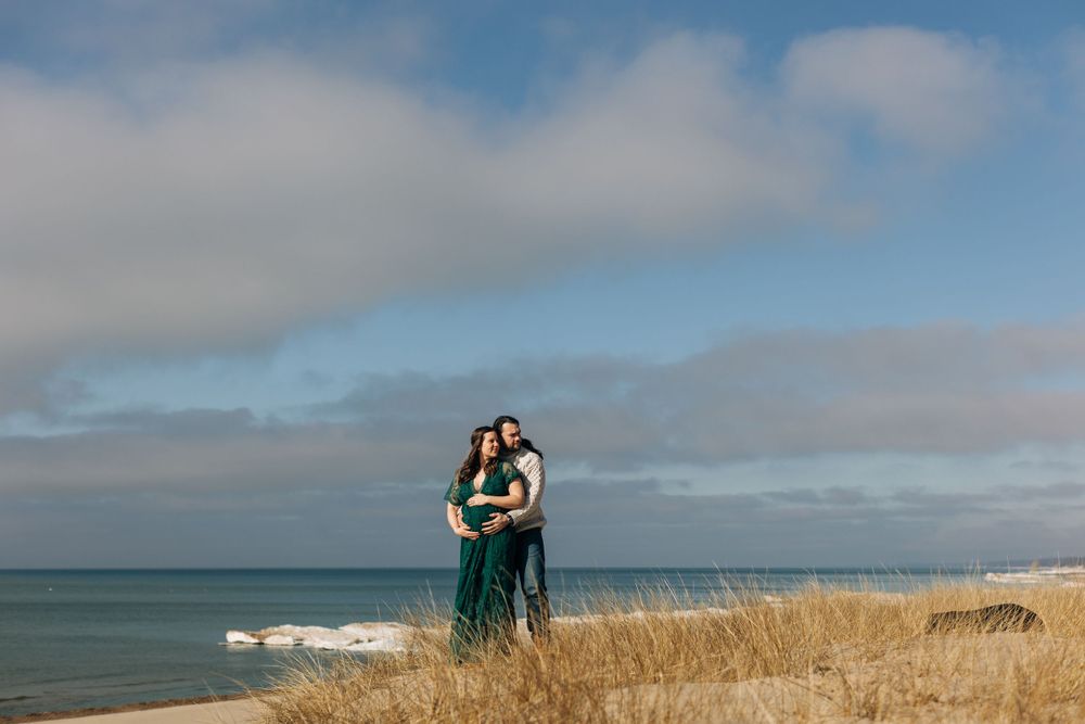 A romantic coastal photoshoot featuring a couple embracing on a grassy dune overlooking the water.