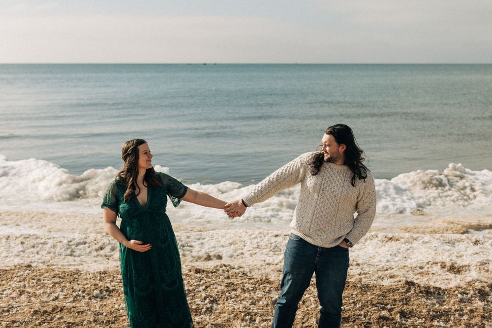 A couple holds hands while standing on a beach with waves crashing behind them.