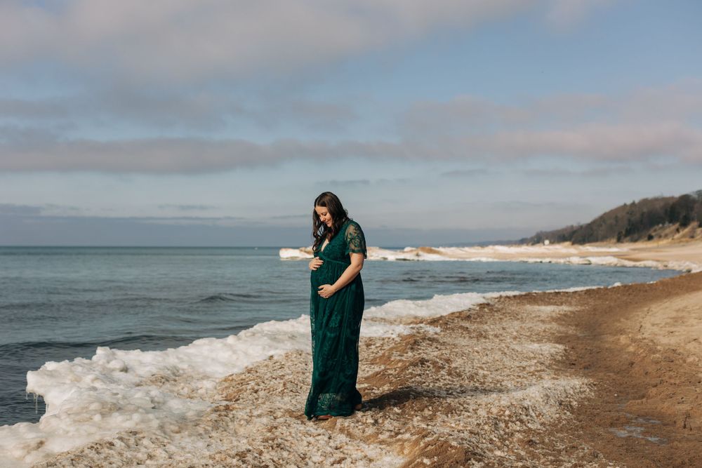 Person in a flowing emerald dress stands alone on a beach looking at the ocean waves.