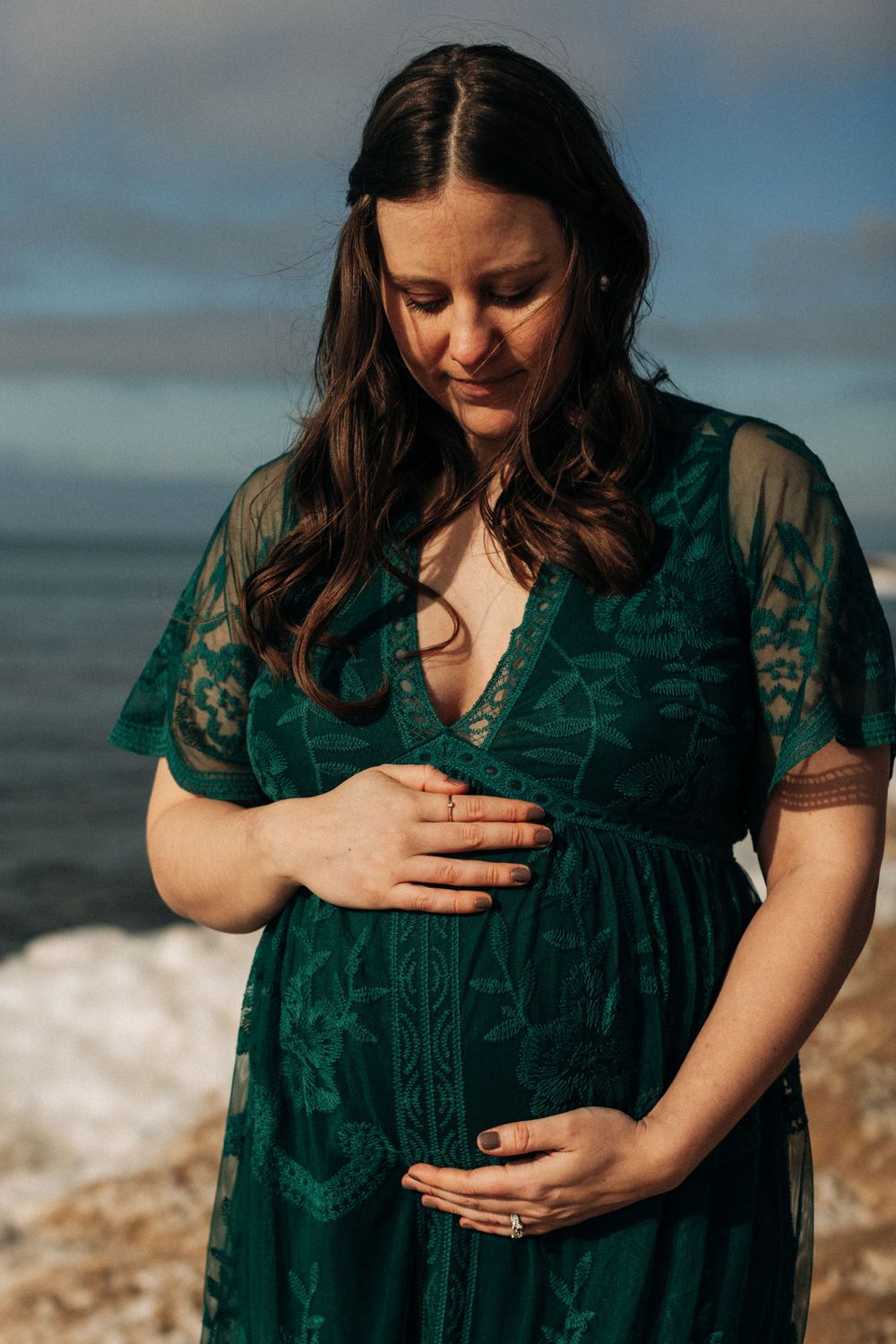 Close-up of person in emerald green lace dress touching their pregnant belly by the ocean.