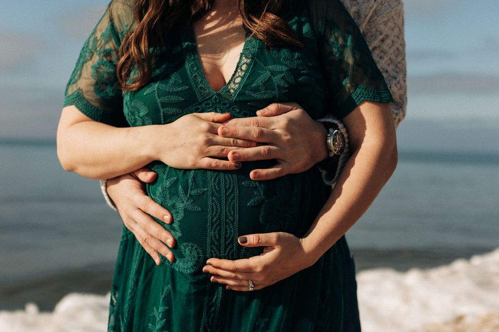 Hands gently cradling a pregnant belly in an emerald green lace dress by the ocean.