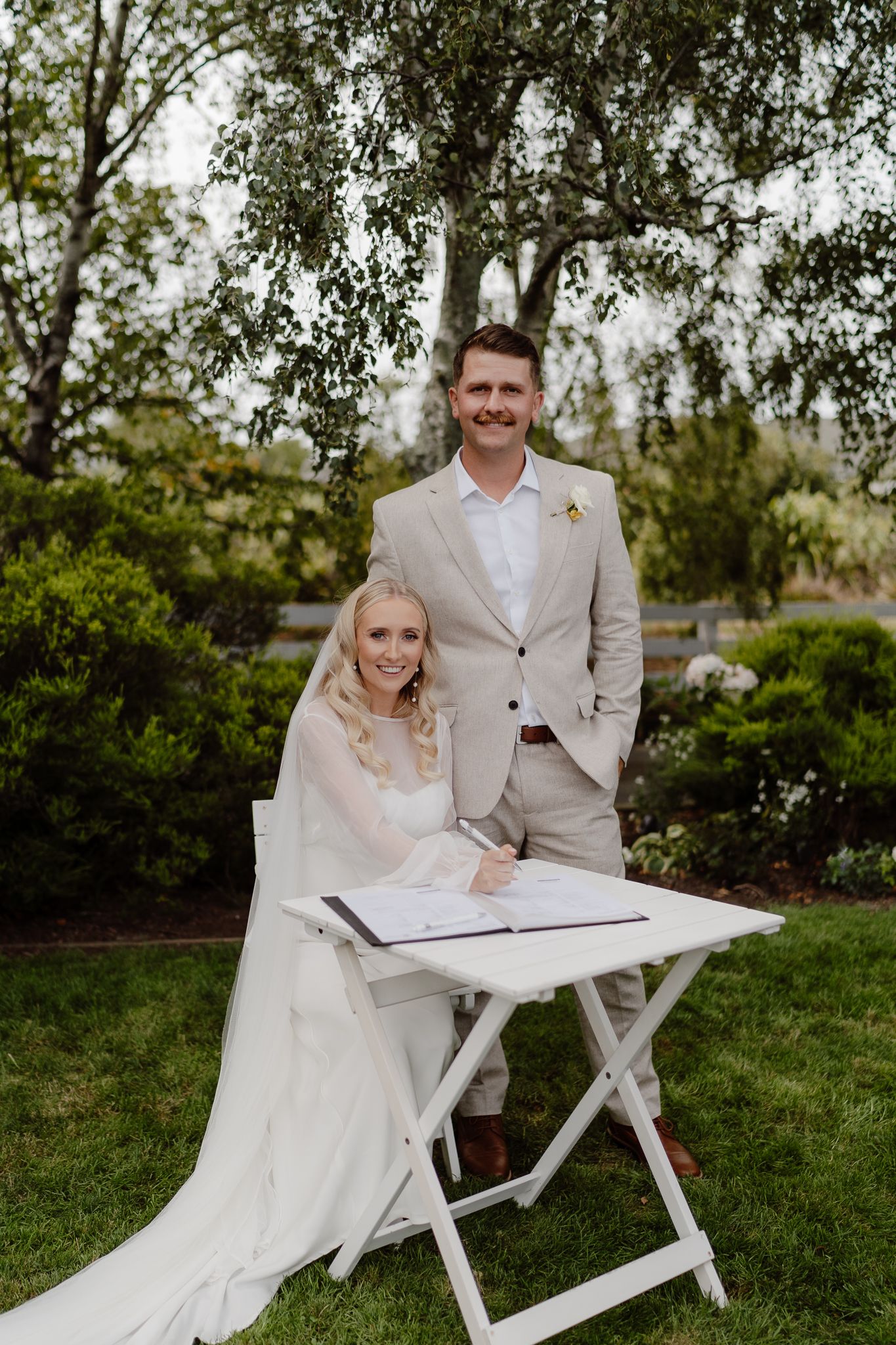 A wedding portrait of a bride sitting and signing her wedding papers and her husband standing beside her