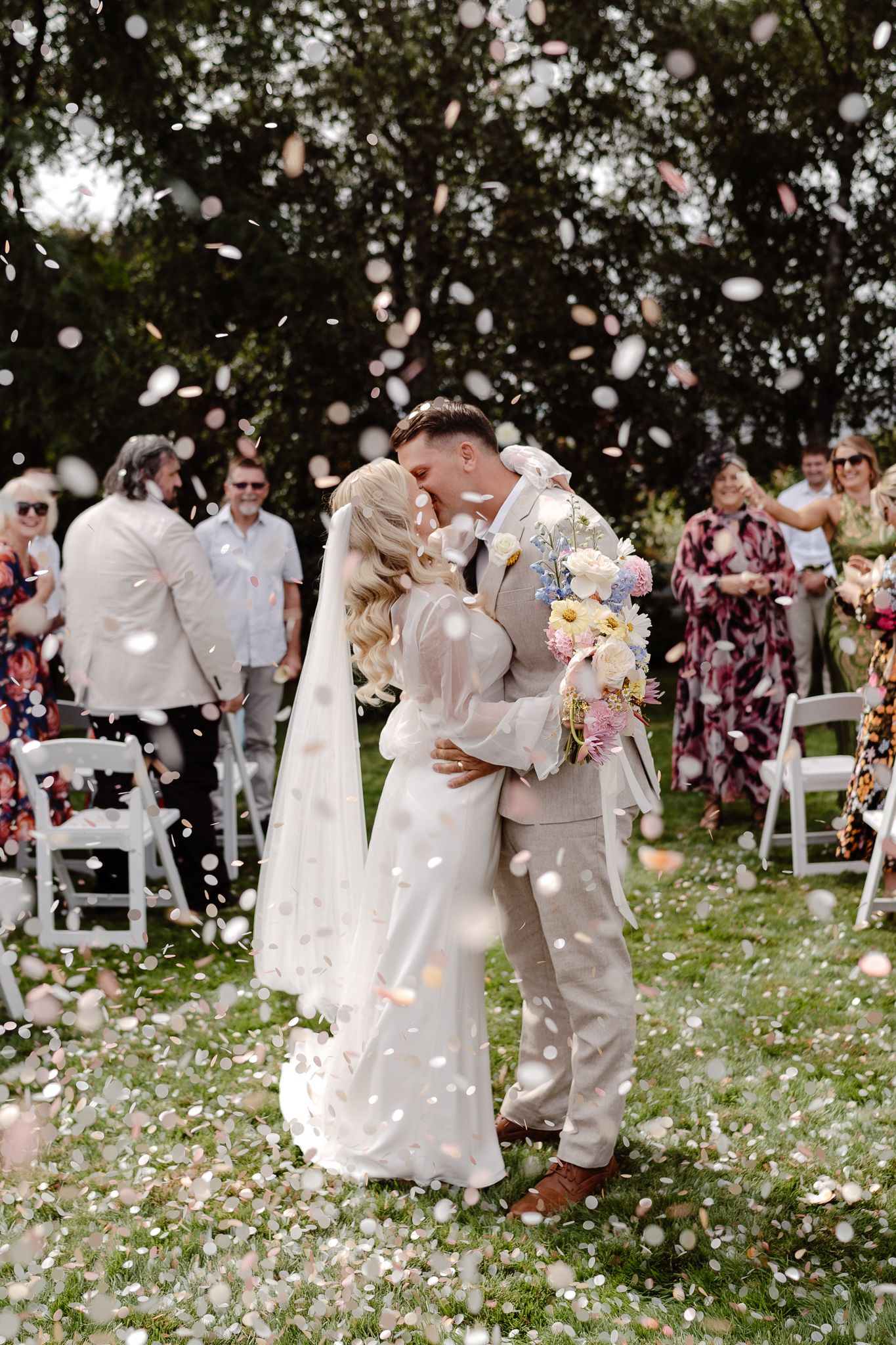 A candid wedding photo of a bride and groom kissing in the wedding aisle while biodegradable confetti is around them