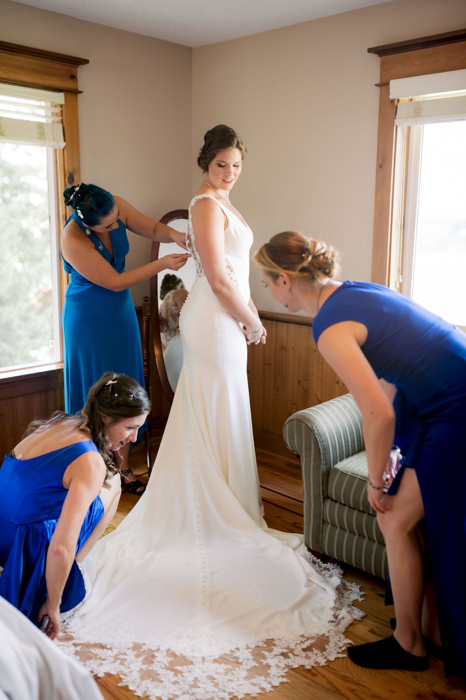 Bridesmaids in blue dresses help arrange a white wedding gown's train in a sunlit room.