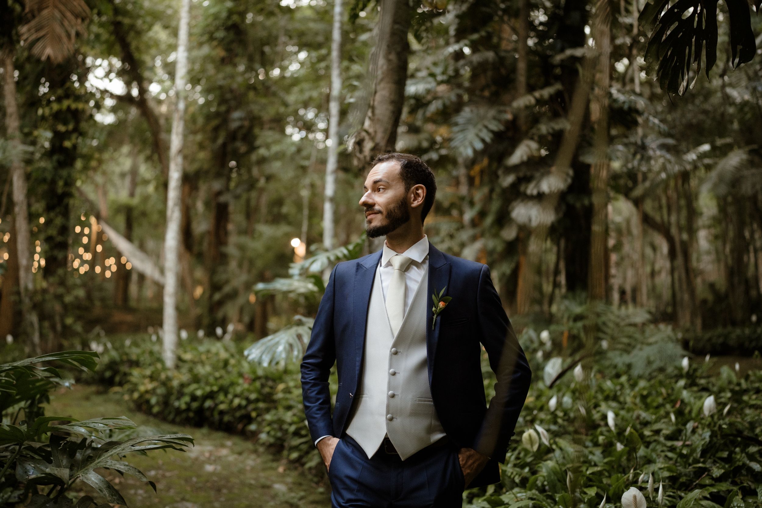 A formal navy suit and cream vest stand out against a lush forest backdrop during golden hour.