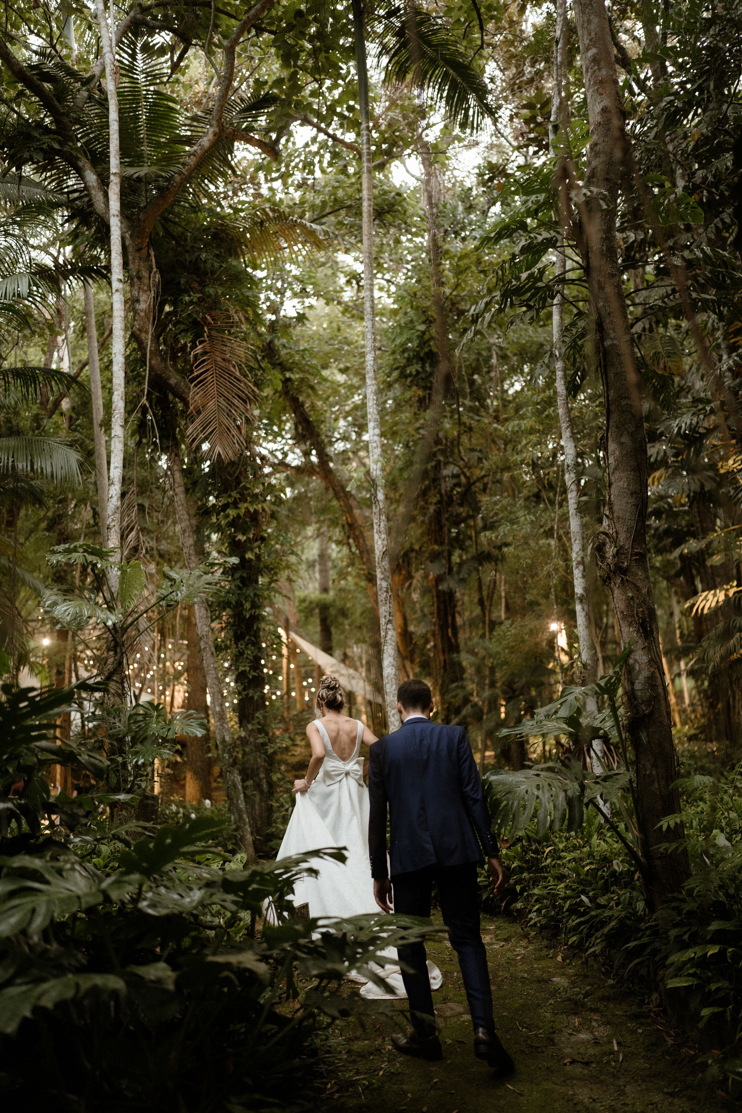 A couple walks hand in hand through a magical forest path lined with tall trees and string lights.