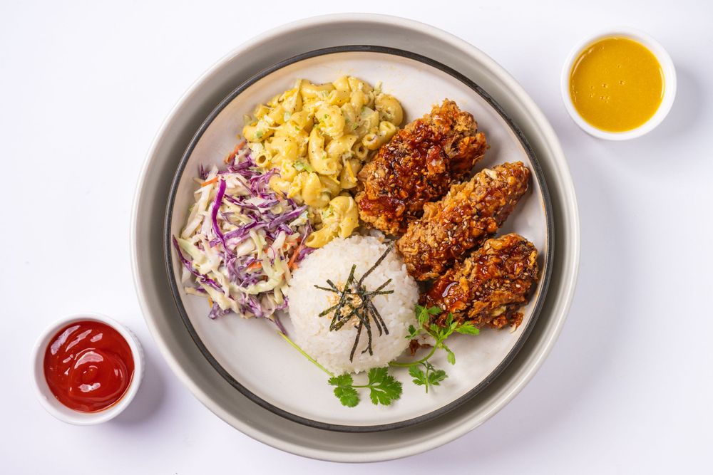 Rice plate with crispy fried chicken, coleslaw, and dipping sauces on gray ceramic dish.