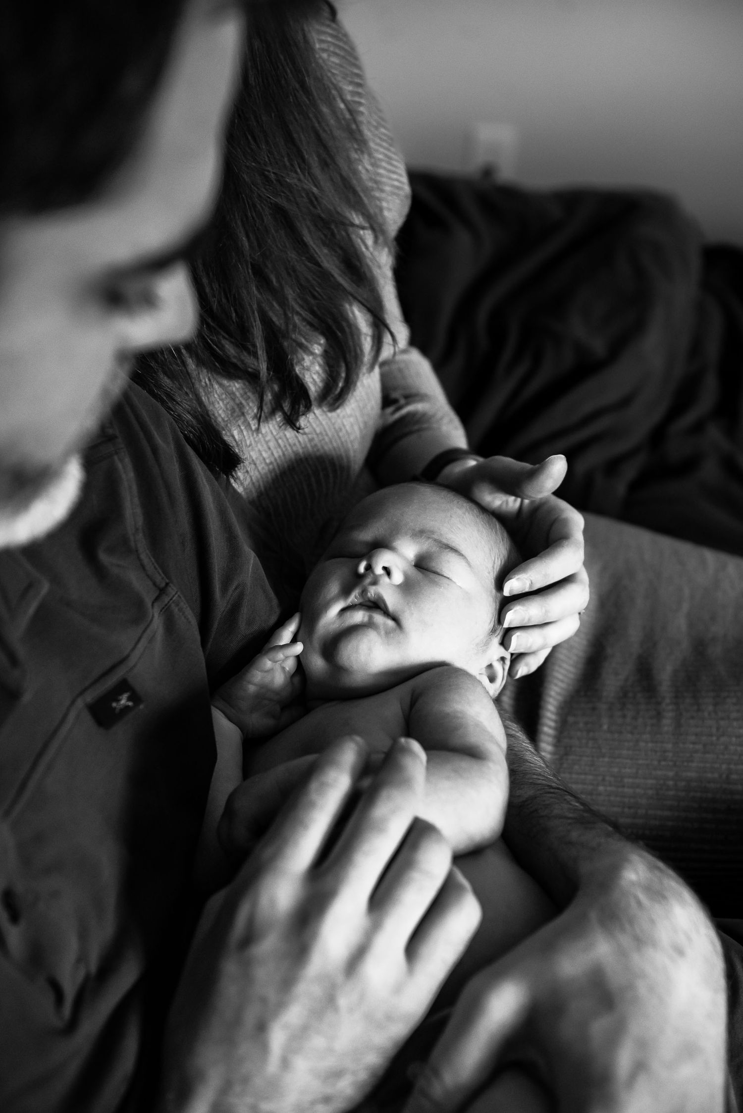 BW image of a newborn baby in the arms of his father, with his mother's hand on his little head and his tiny baby hand.