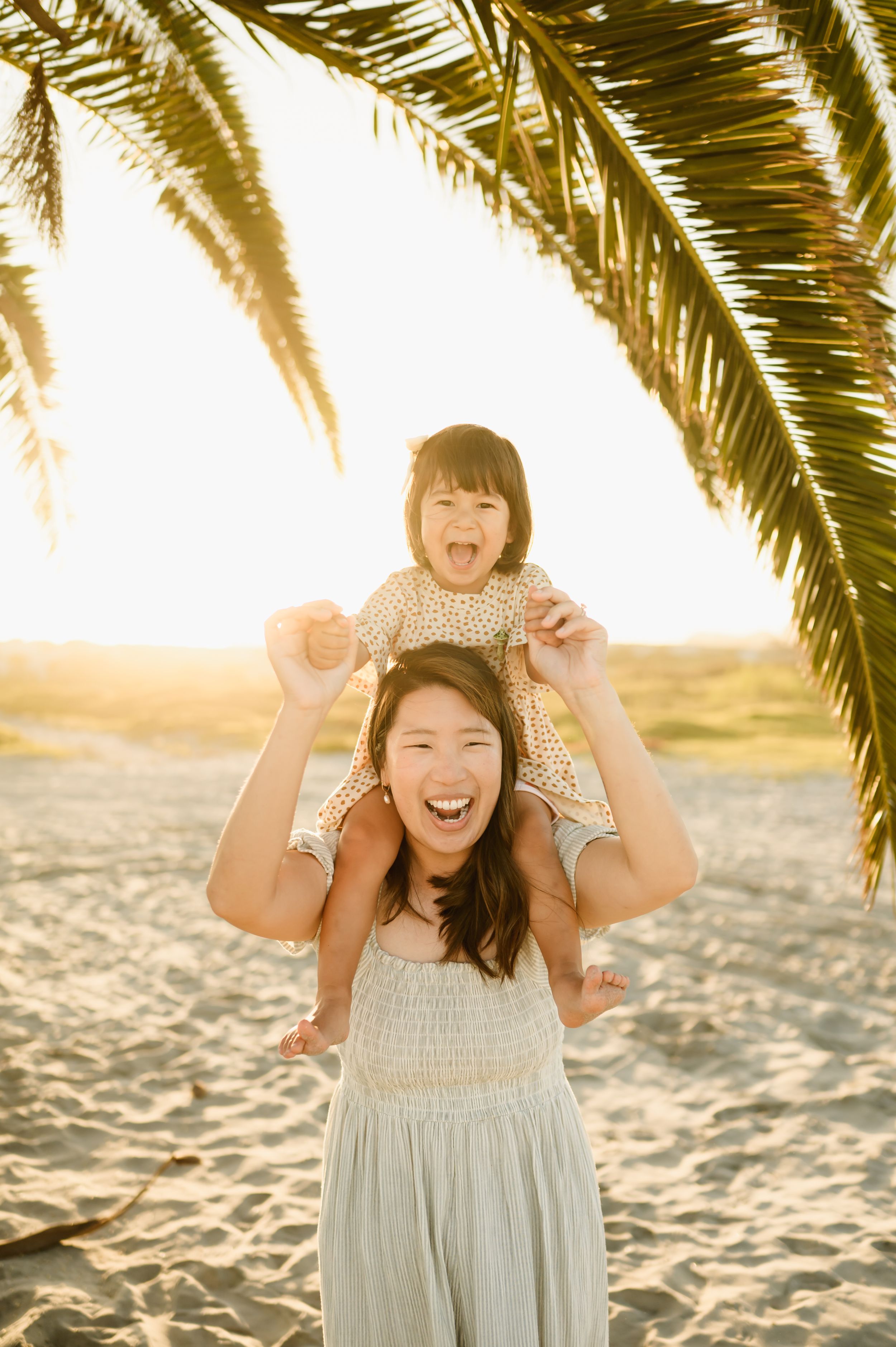 Mom carrying a happy child on her shoulders under a palm tree at Zachari Dunes beach during Ventura family photoshoot