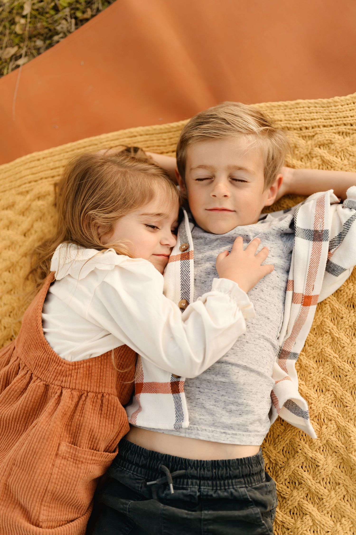 Two children lying on a blanket outdoors, with the younger child resting on the older child's chest.