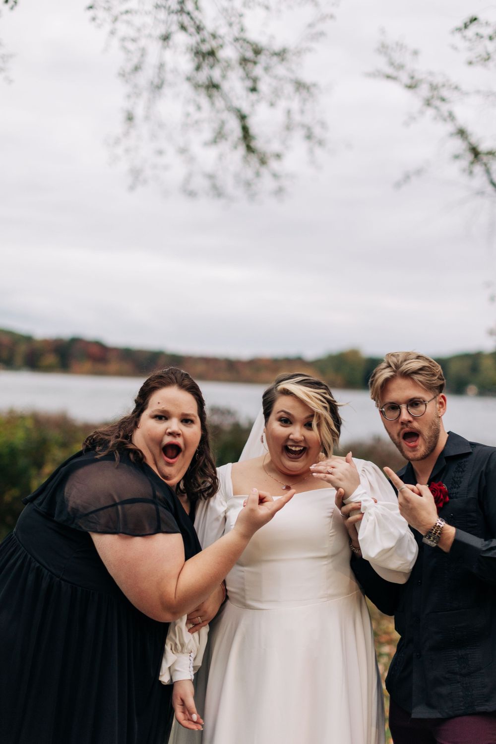 A wedding party reacts with excitement while posing outdoors near a lake on an overcast day.