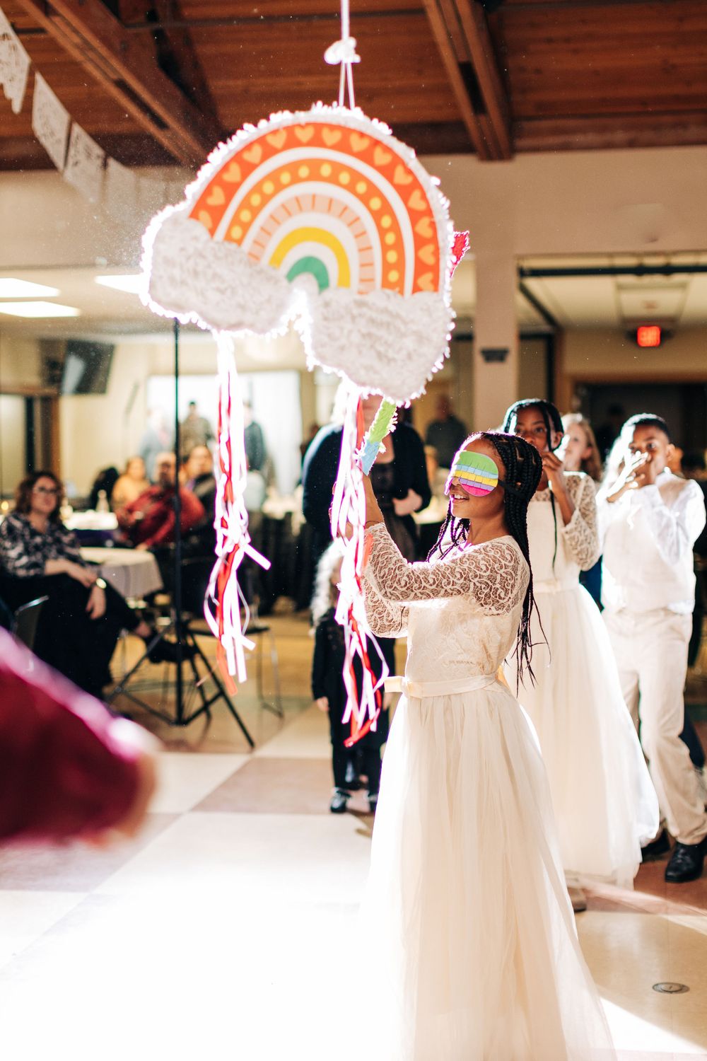 A colorful rainbow piñata hangs from the ceiling at a wedding reception while guests celebrate below.