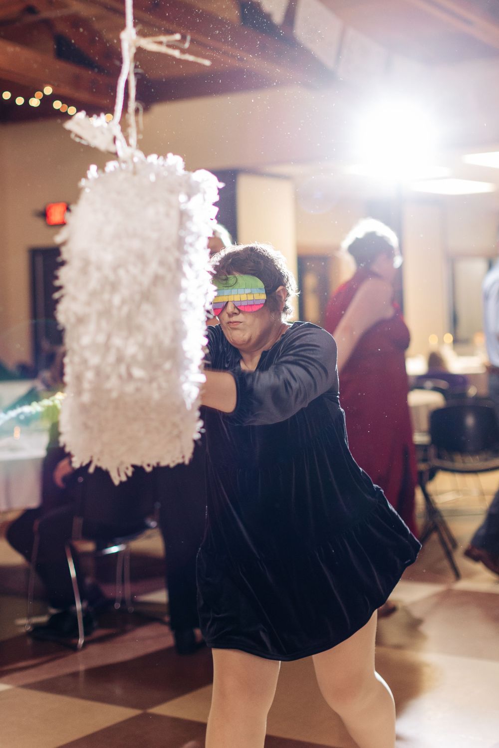 A person wearing neon glasses swings at a white piñata during an indoor celebration.