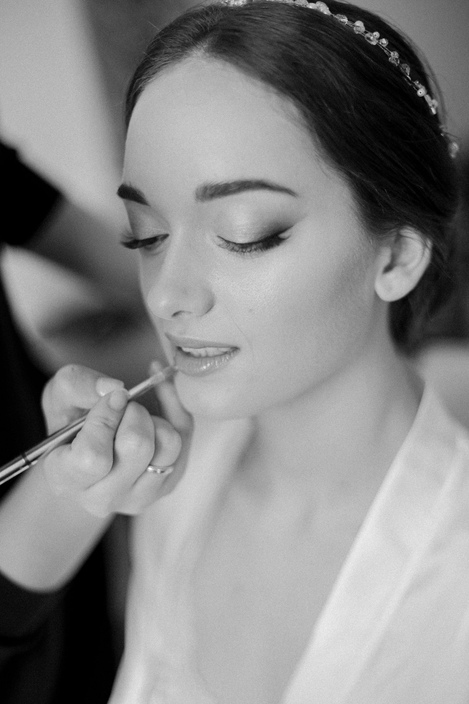 Close-up black and white photo of bride getting professional makeup applied while wearing delicate headpiece.