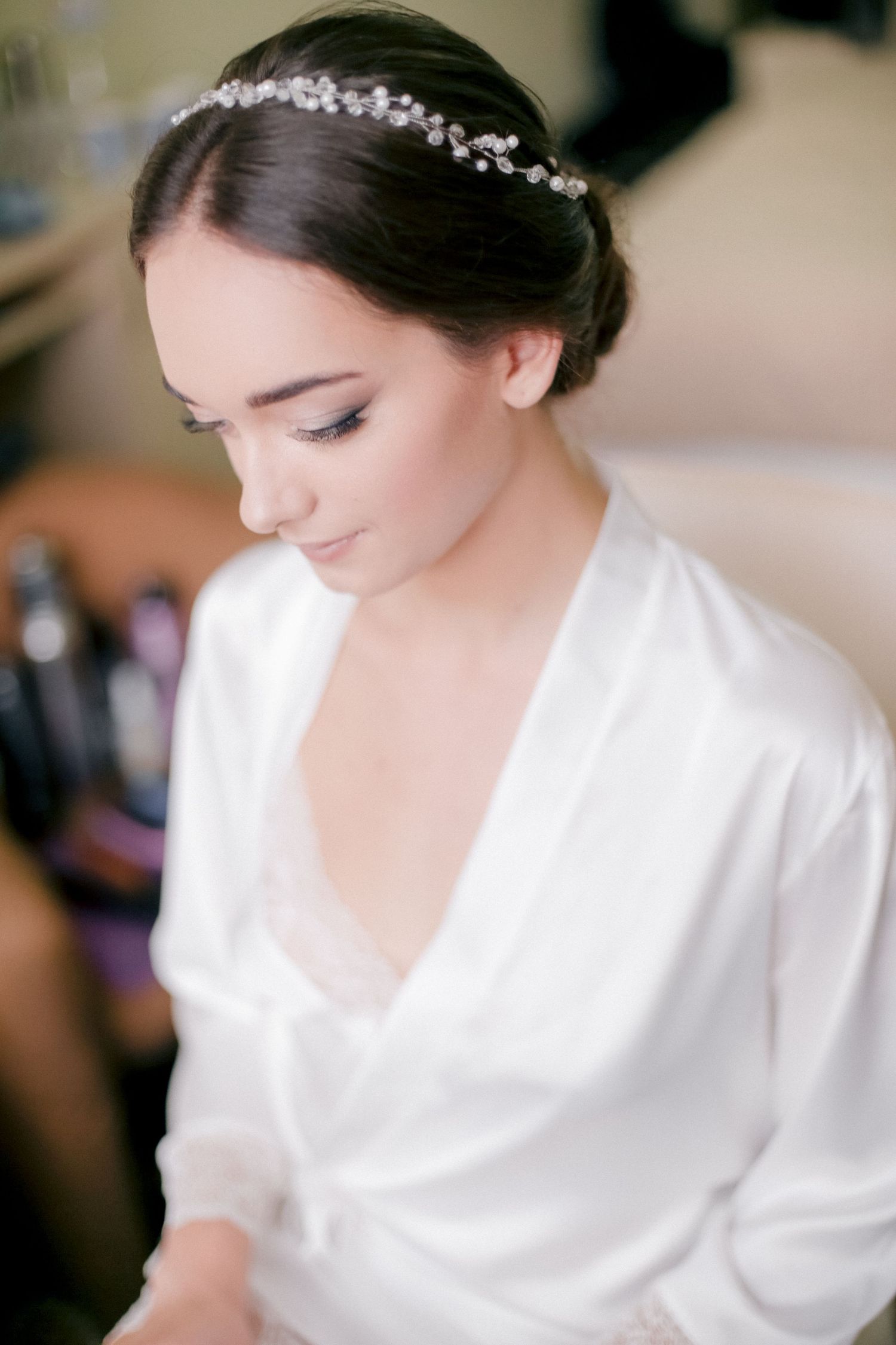 Bride in white robe and crystal headpiece looks down during wedding preparations.