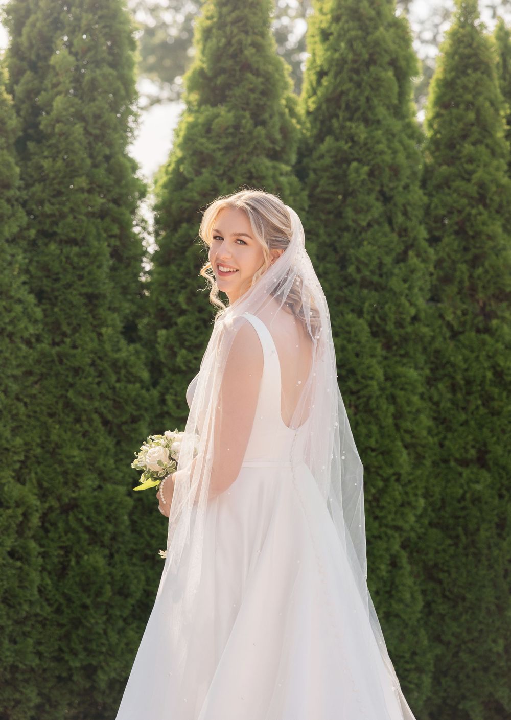 A wedding dress and veil photographed against a backdrop of green cypress trees.