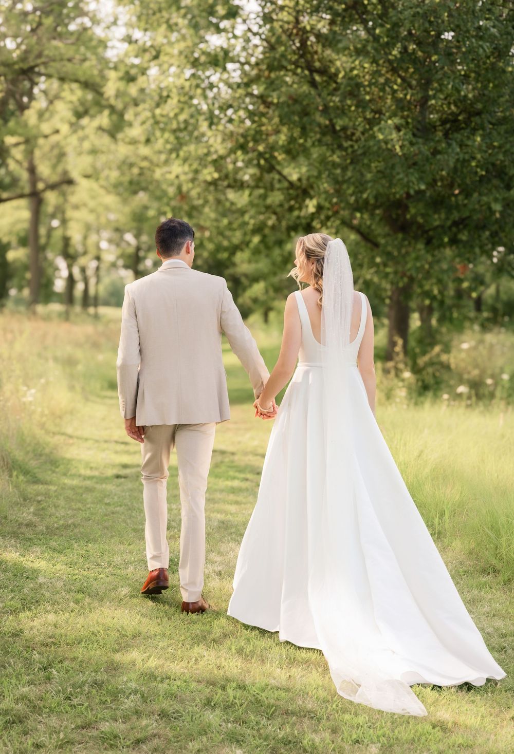 A couple walking through a sunlit meadow in wedding attire.