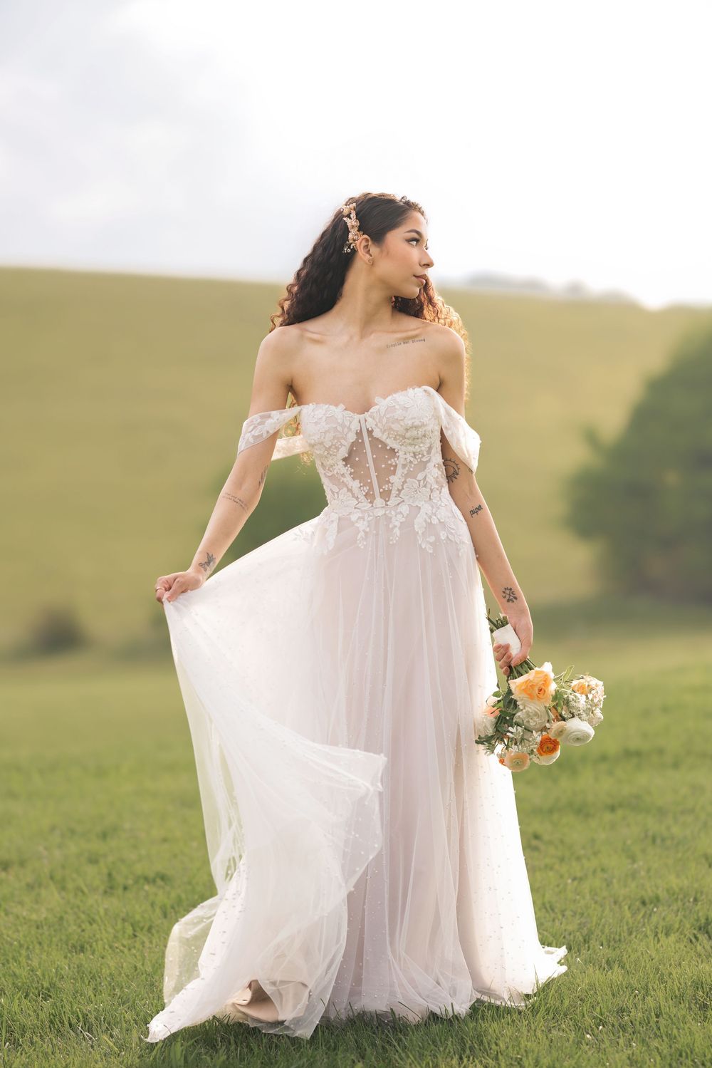A bride in a flowing white off-shoulder gown twirls gracefully in a sunlit meadow holding a small bouquet.