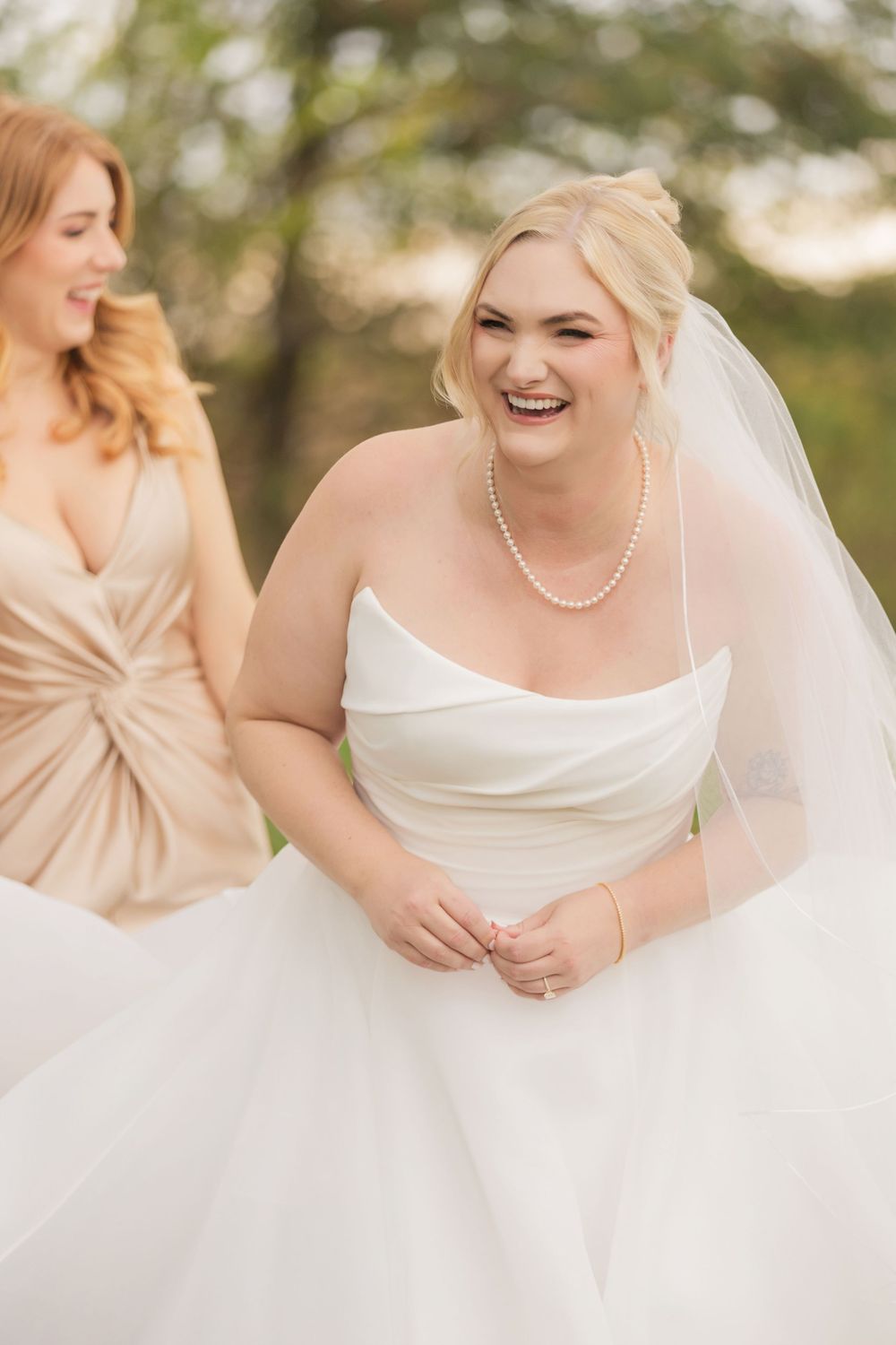A bride in a classic white strapless gown and pearl necklace shares a joyful moment with bridesmaid in champagne dress outdoors.