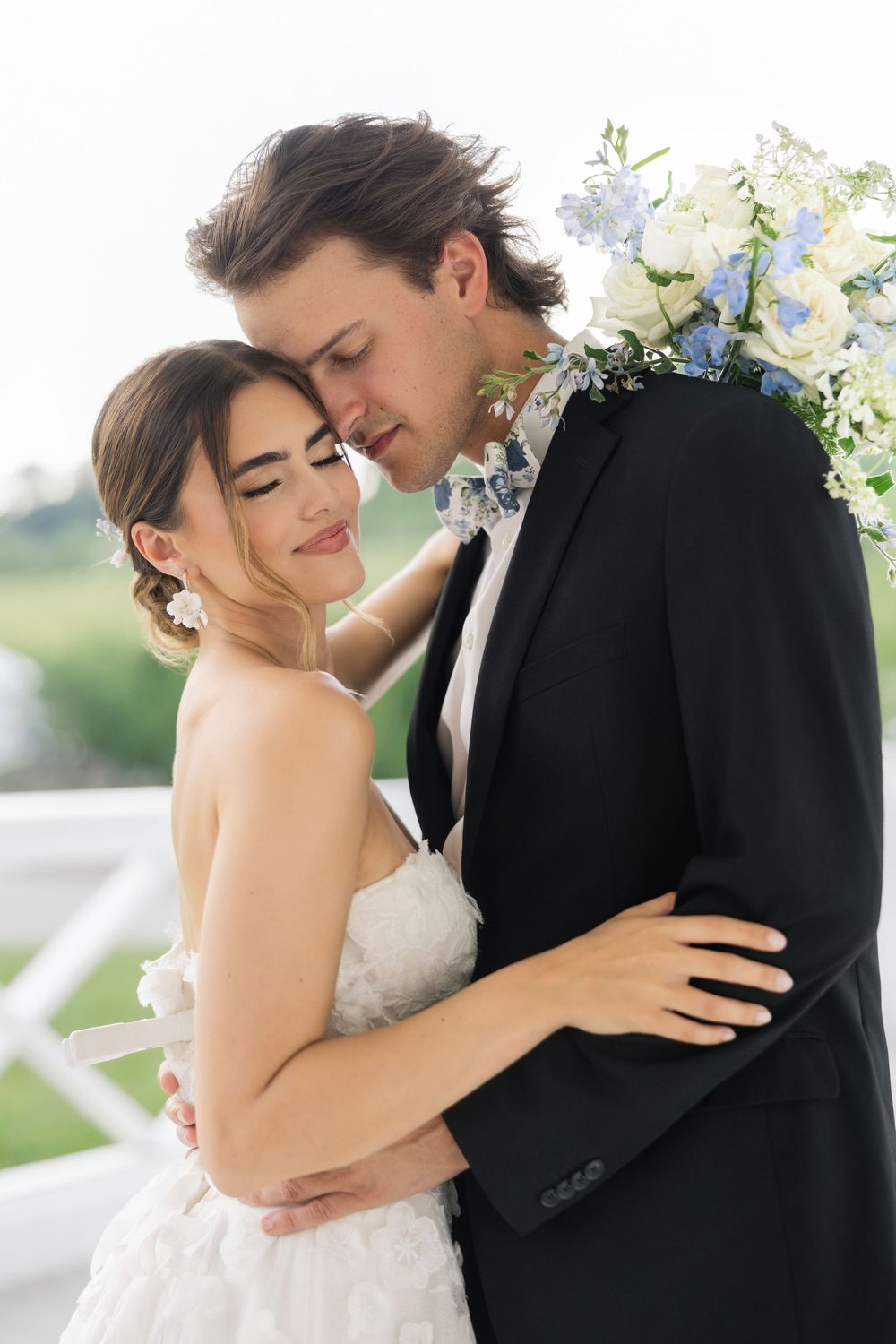 A romantic wedding couple embraces in an intimate moment surrounded by white floral decorations outdoors.