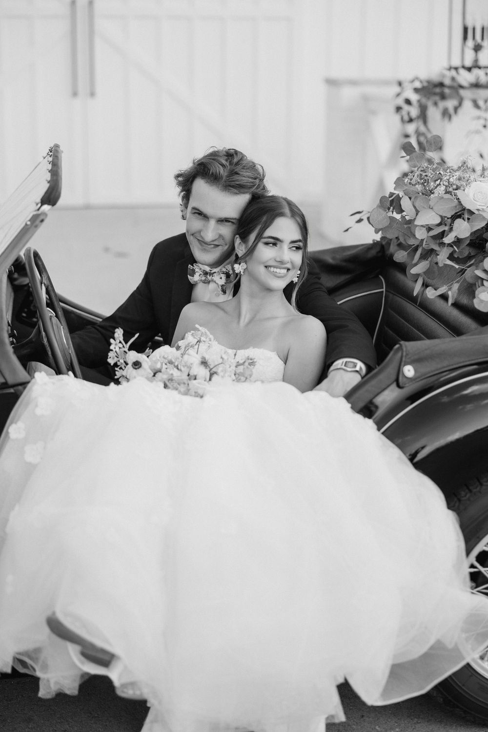 A black and white wedding photo of newlyweds sitting in a vintage car with a flowing white wedding dress.