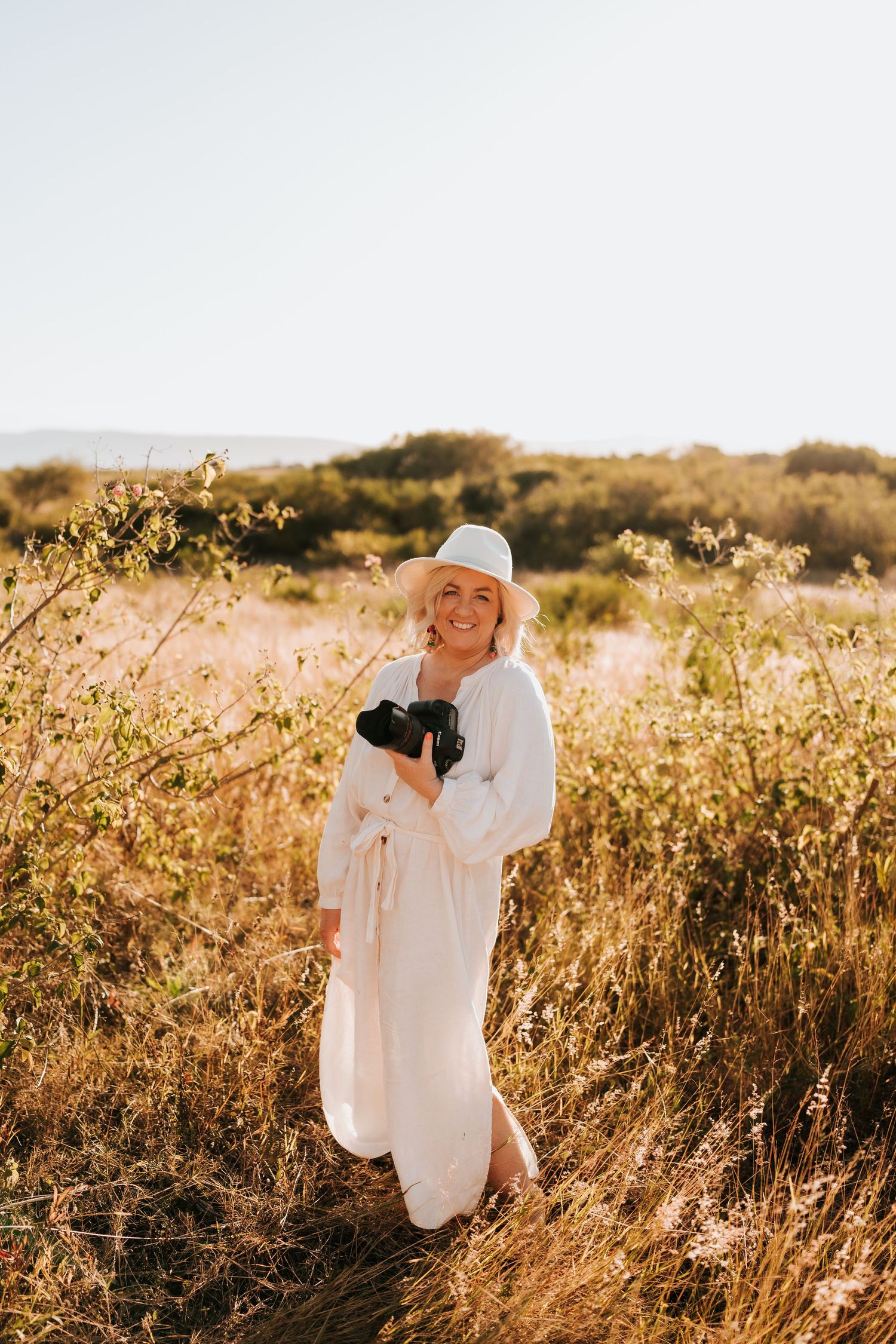 Photographer in flowing white dress and hat poses with camera in golden sunlit field.