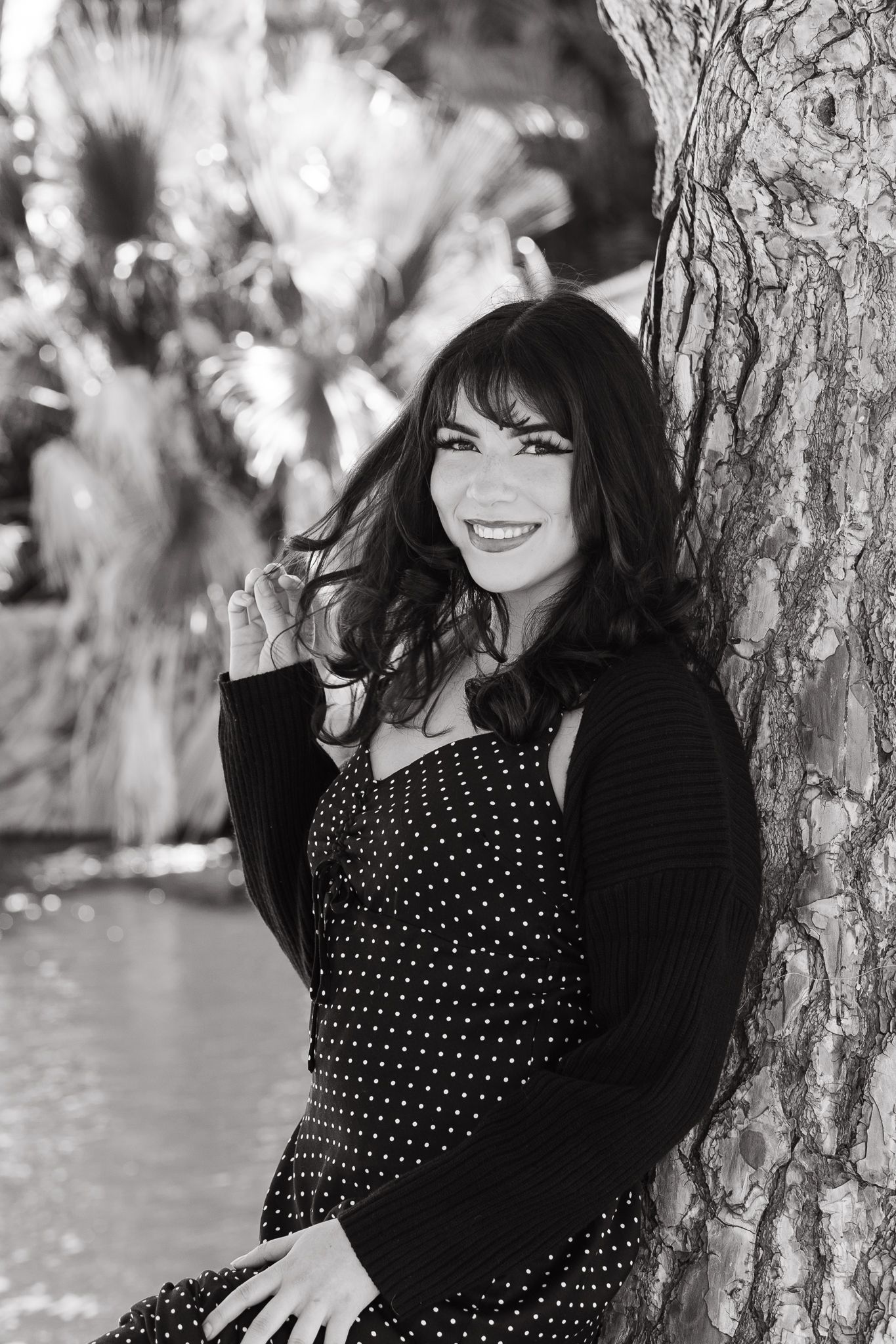 A black and white portrait of someone smiling while sitting against a tree trunk.