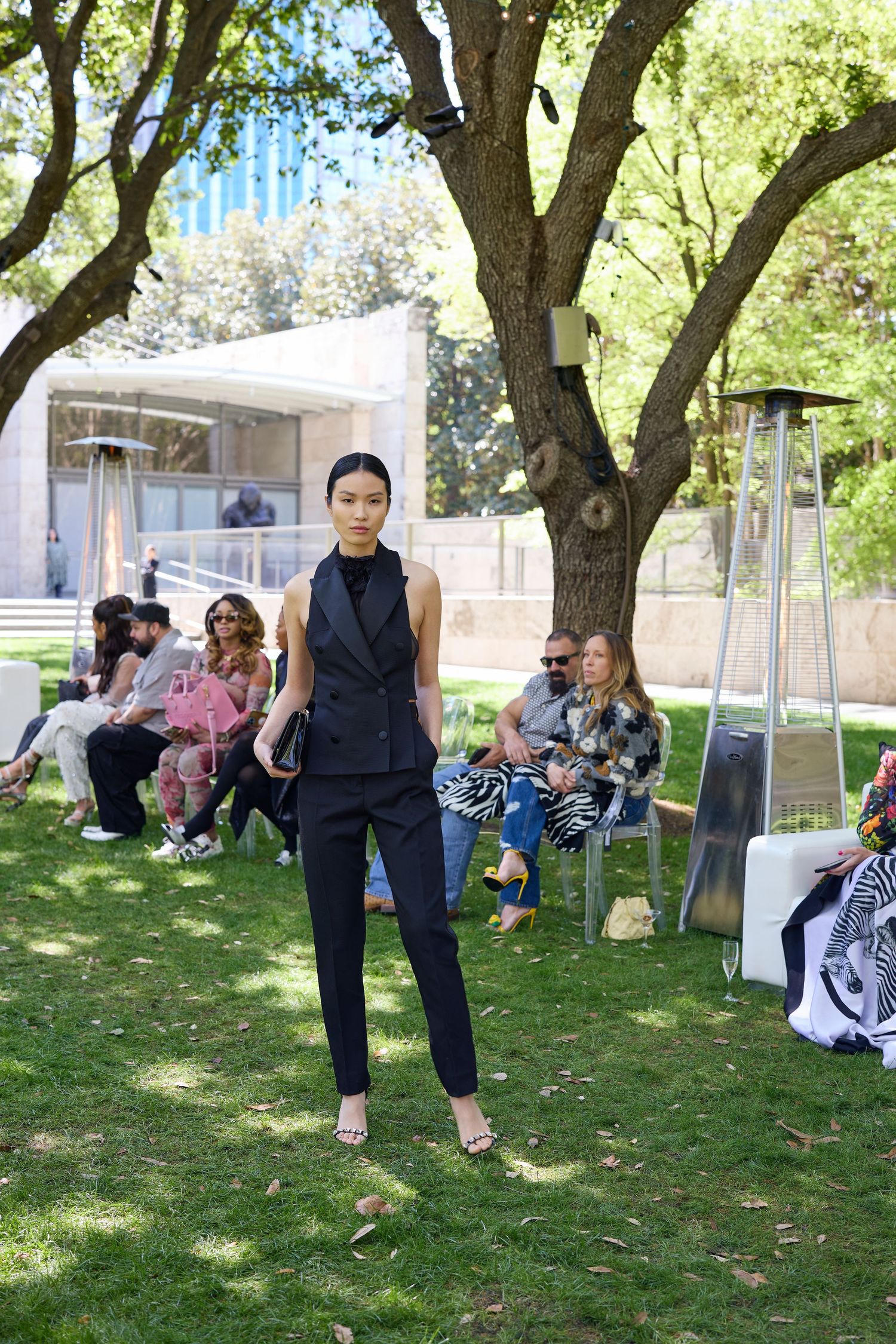 Fashion models showcase designer outfits at an outdoor garden runway show with spectators seated under trees.