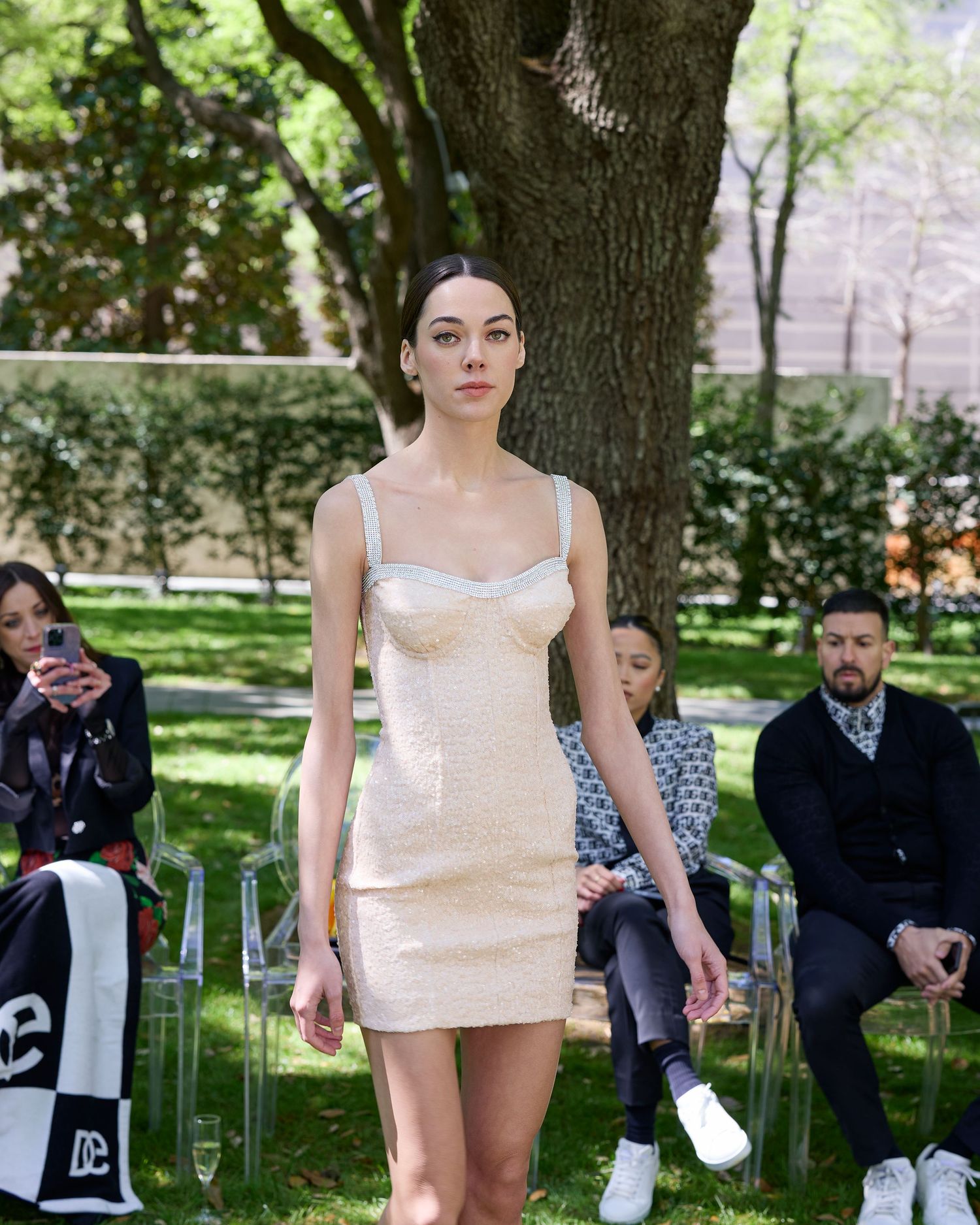 A pale pink fitted mini dress is presented during an outdoor fashion show with attendees seated on white chairs.