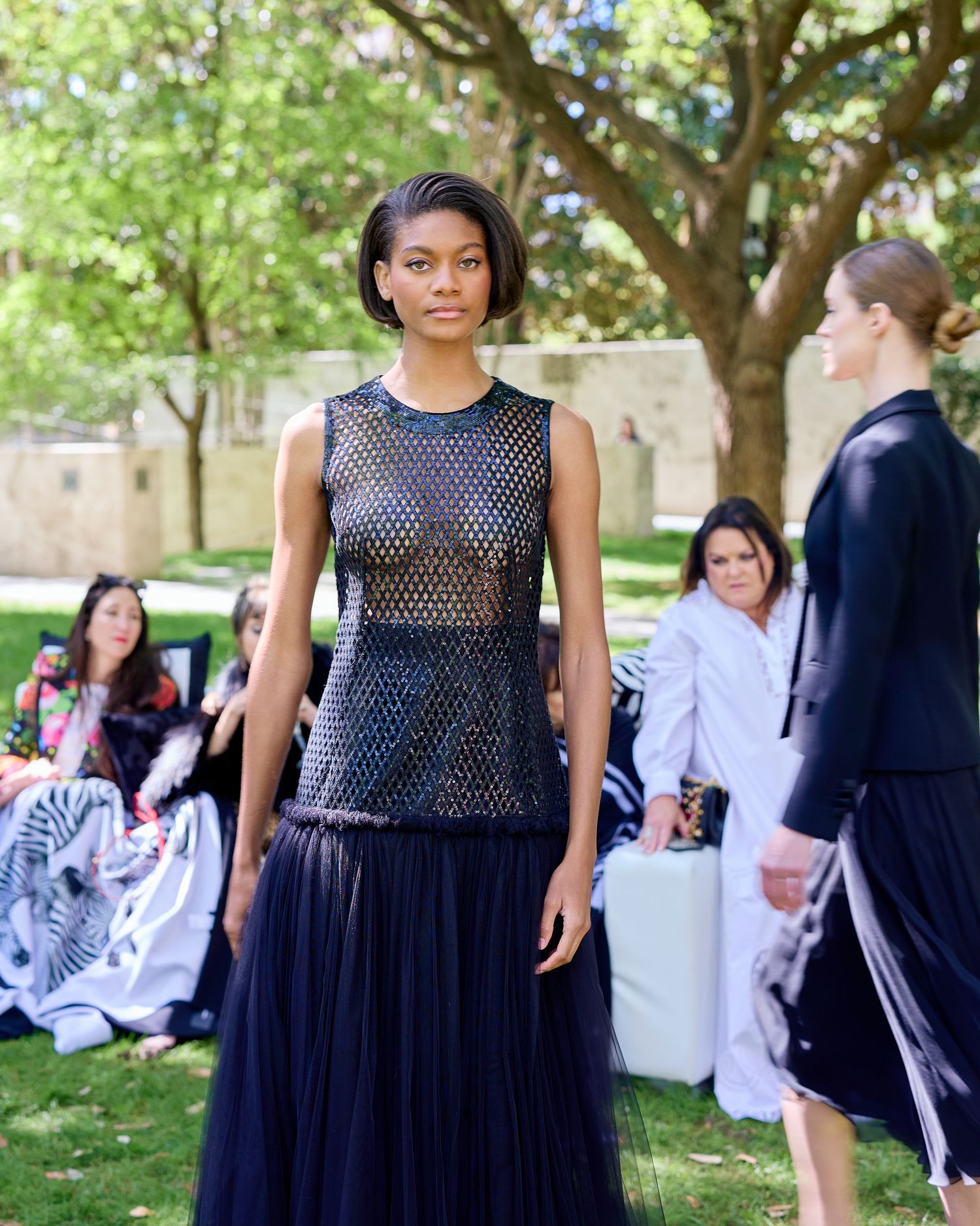 A navy blue evening gown with mesh detailing and pleated skirt is displayed at an outdoor fashion presentation.