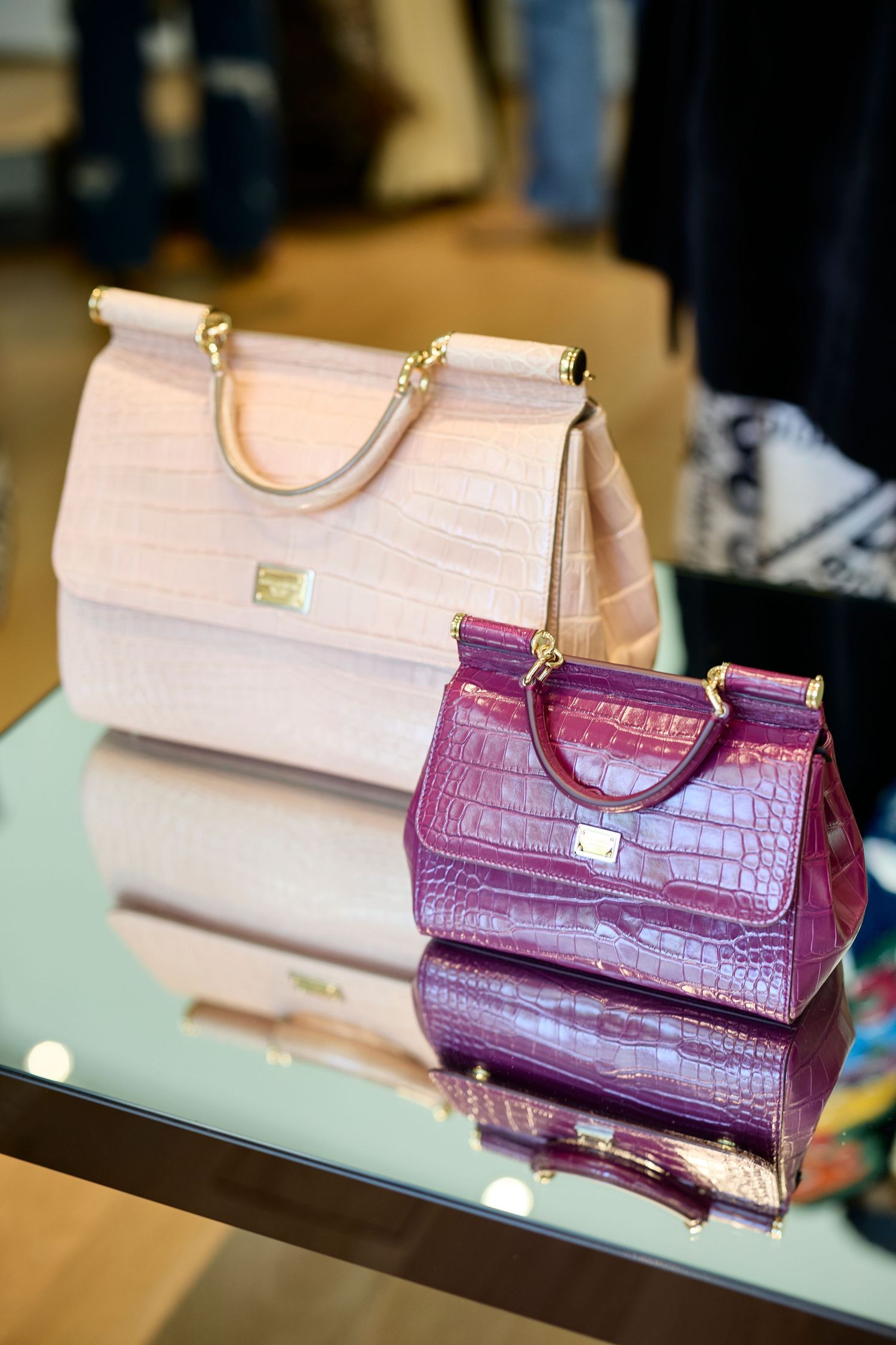 Luxurious crocodile-embossed handbags in pink and purple displayed on a glass surface.