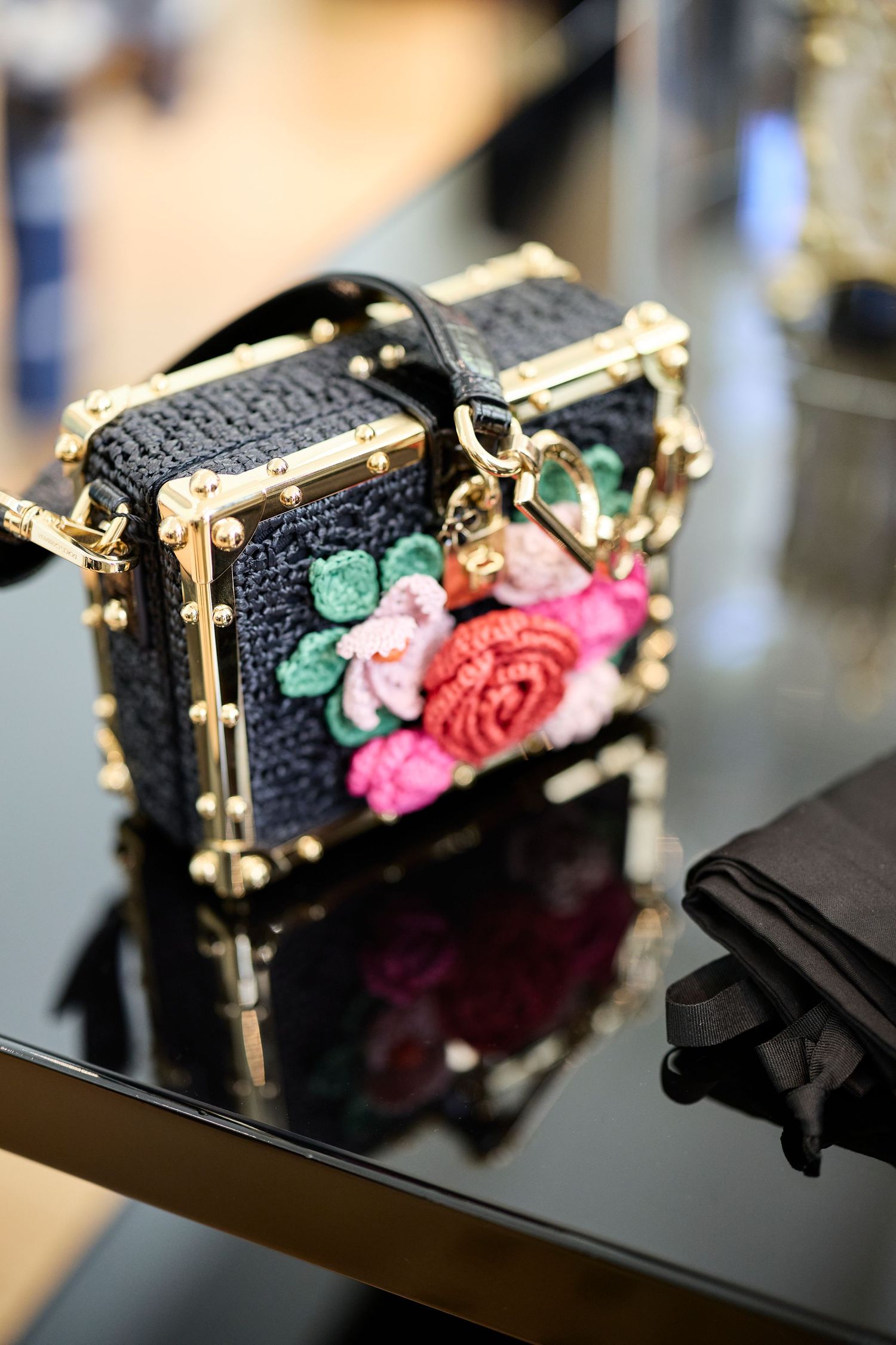 Black embellished evening bag with floral details and gold hardware displayed on reflective surface.
