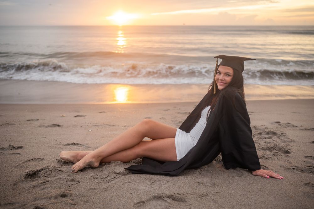 Senior girl graduation photoshoot session in cap and gown sitting in sand at beach sunset Melbourne Cocoa Vero Florida Brevard County