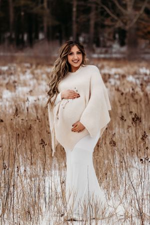 Winter maternity photos of woman in white dress standing among dried reeds in snowy field.