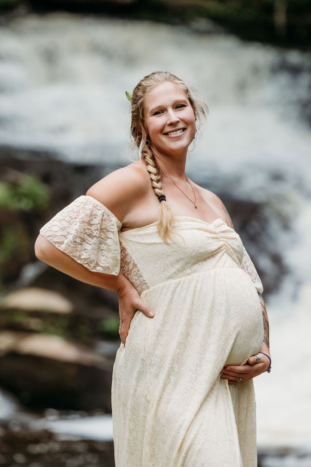 Woman in a flowing cream lace dress poses by a waterfall.