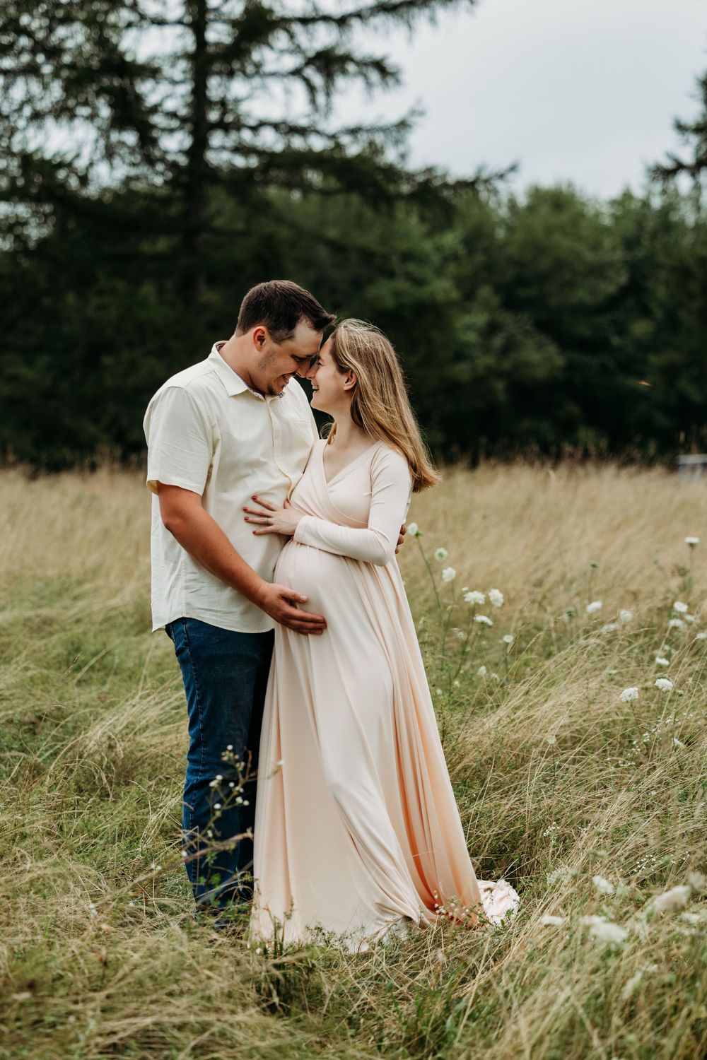 Romantic couple standing together embracing pregnancy in a grassy meadow with evergreen trees in the background.