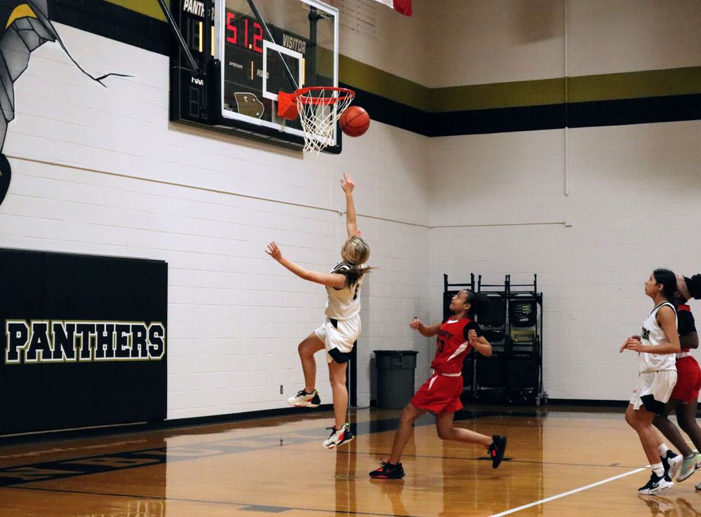 Player in white uniform jumps to score during basketball game against team in red.