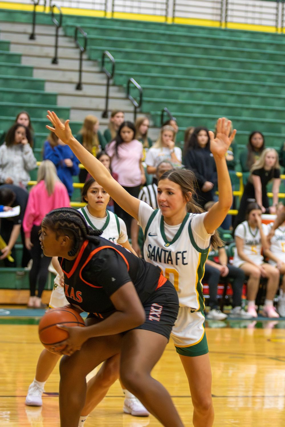 Basketball players compete for position while spectators watch from green bleachers in background.