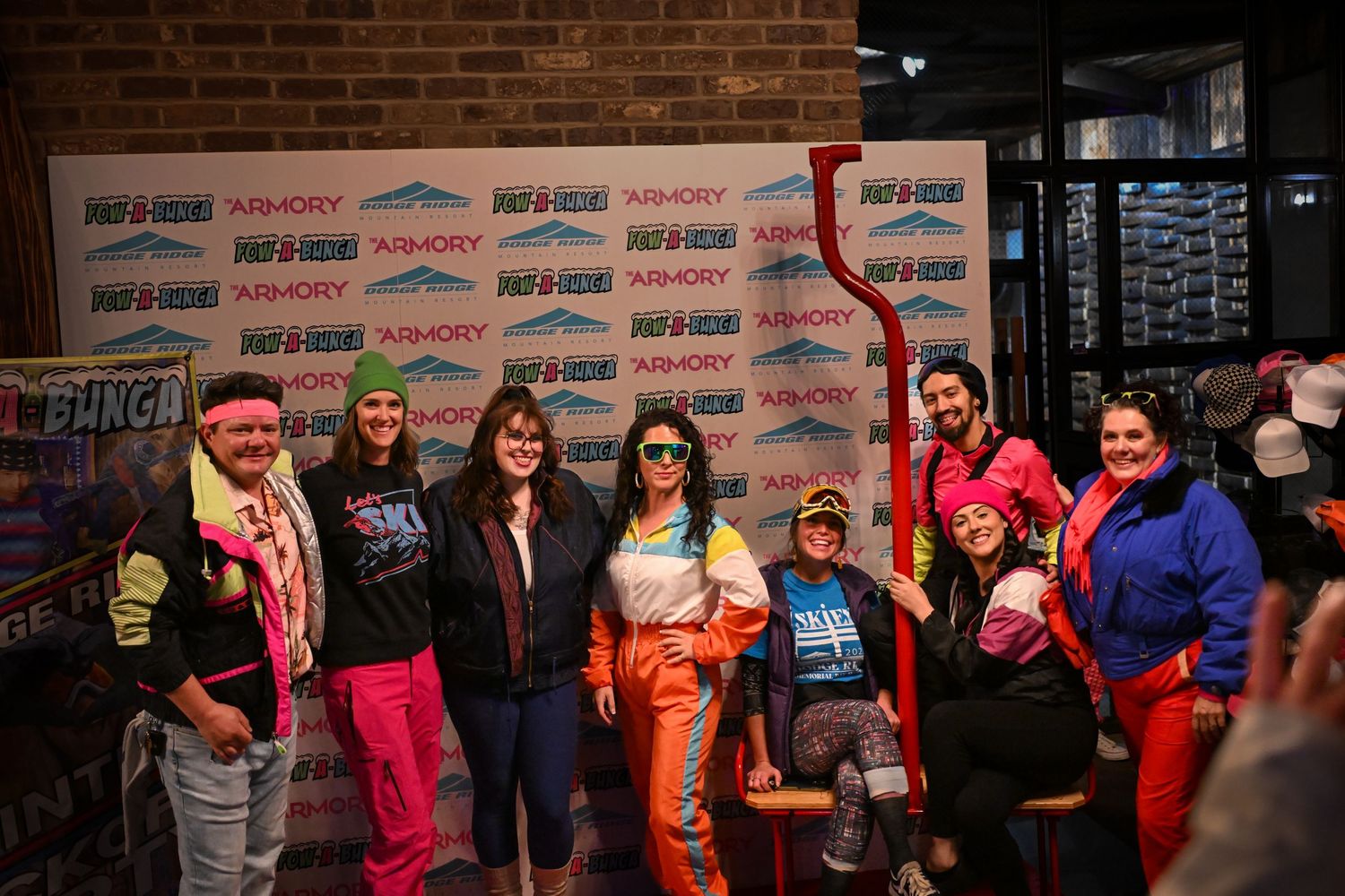 Group of people in colorful costumes and outfits pose together against a brick wall backdrop.