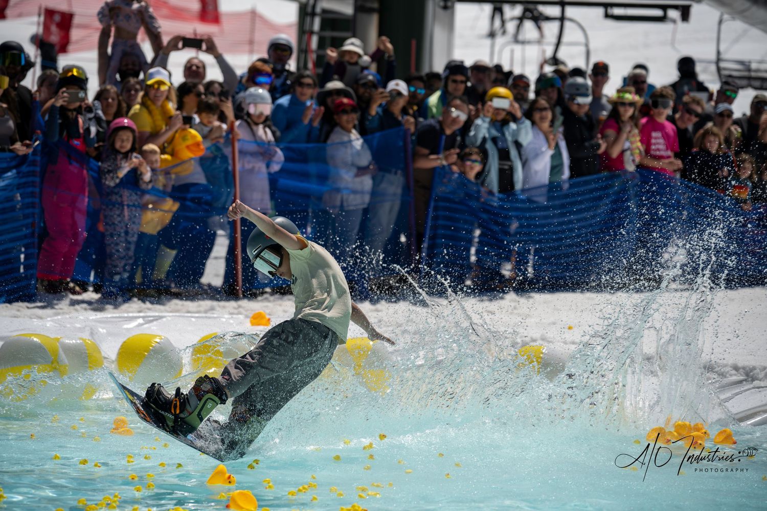 Skier splashes through pool of water during pond skimming competition with large crowd watching.
